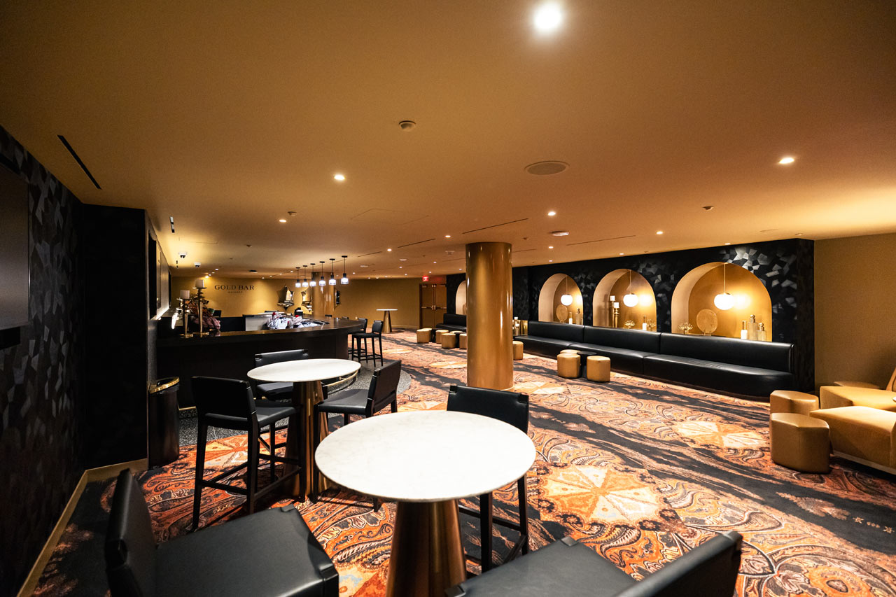 A stylish lounge area featuring a bar labeled "Gold Bar," black and gold seating, marble tables, and arched alcoves with warm lighting and decorative elements.