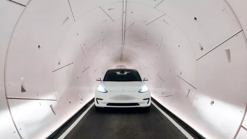 A head-on, centered view of a white Tesla Model 3 with its headlights on, parked inside a brightly lit, narrow, white cylindrical concrete tunnel.