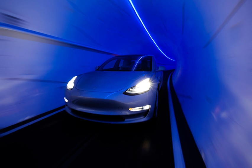 A white Tesla Model 3 driving through a narrow, curved tunnel illuminated entirely by vibrant blue neon lighting, creating a dramatic motion blur effect.