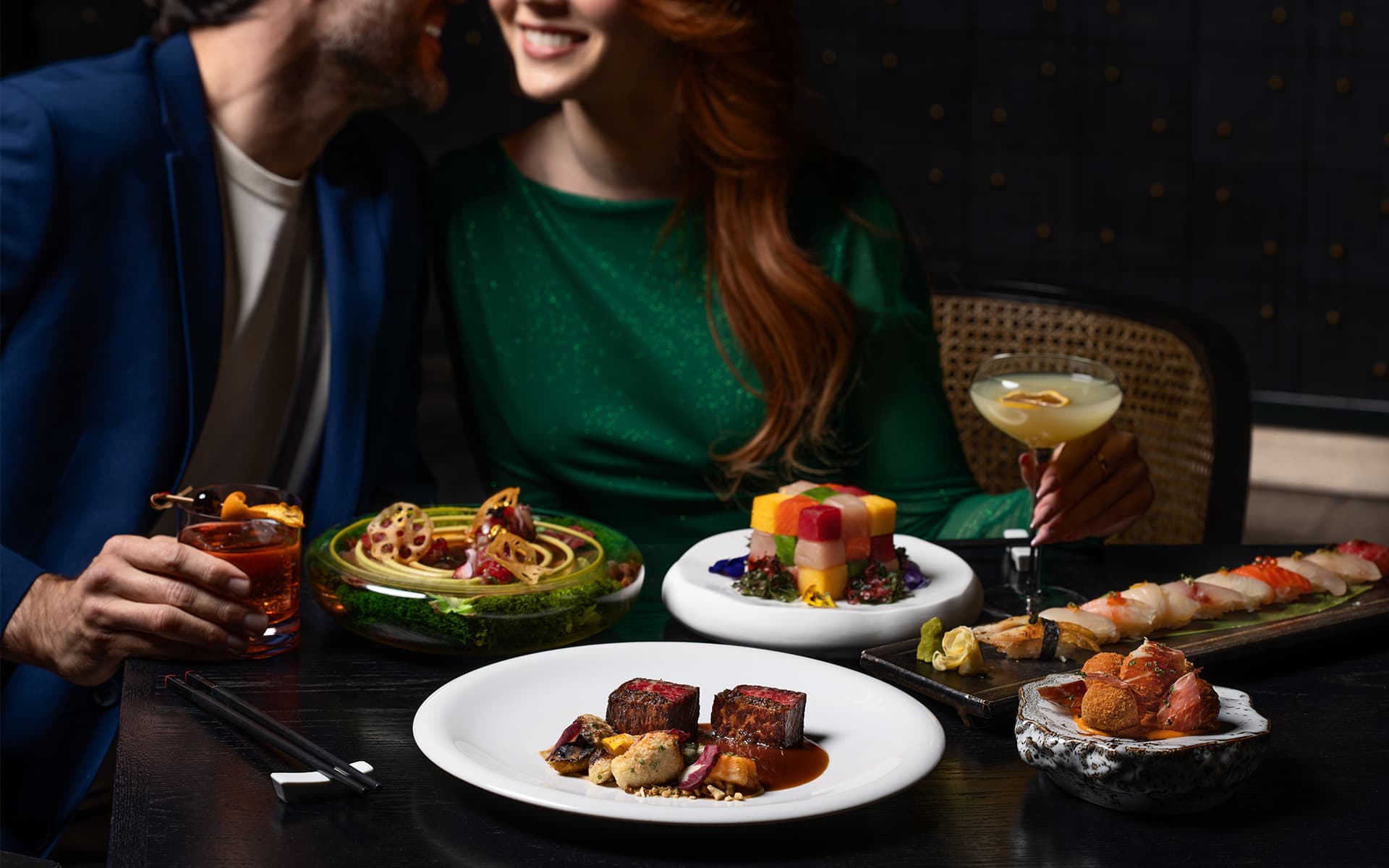 Couple enjoying a fine dining meal with sushi, steak, plated dishes, and cocktails at a restaurant table.
