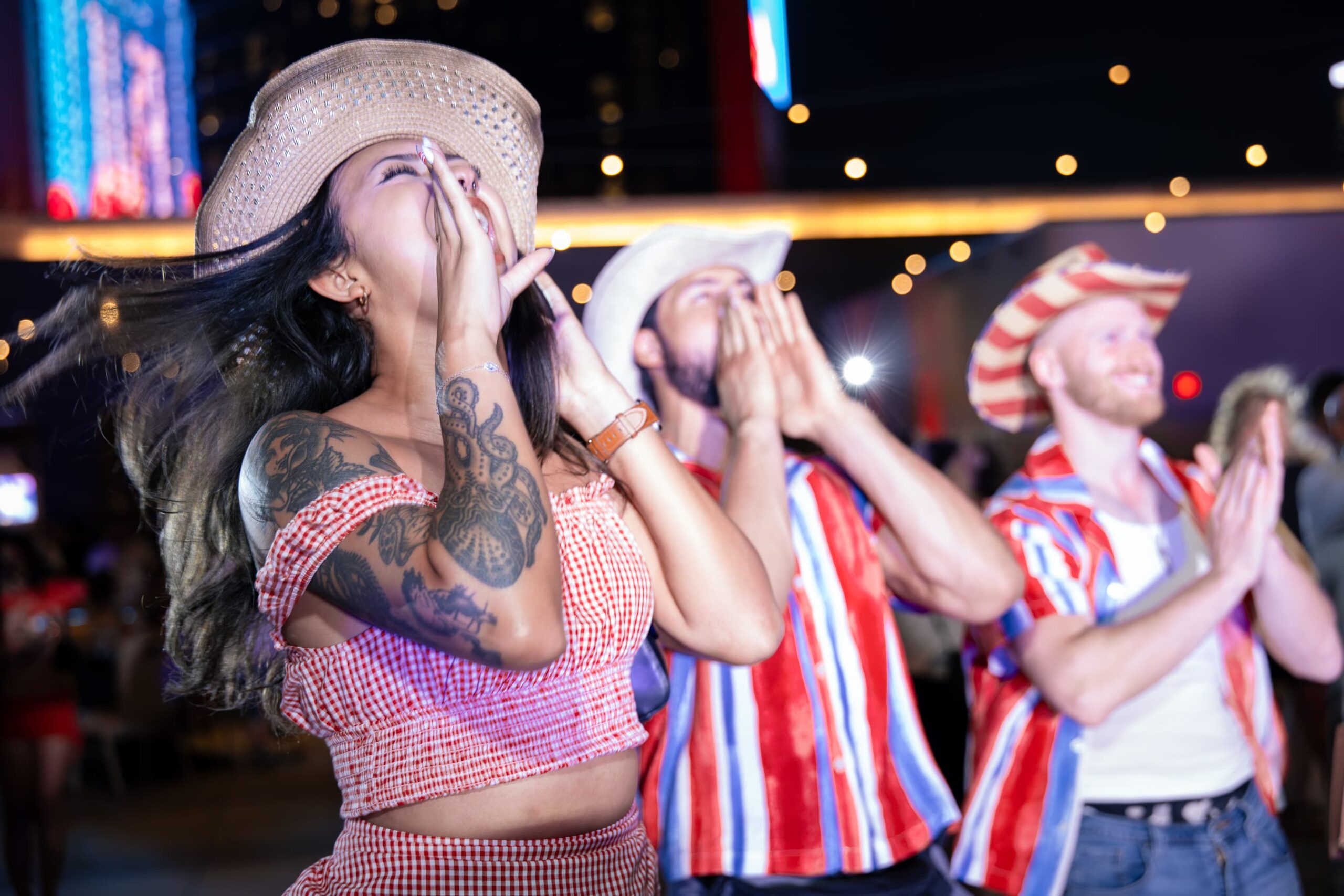 Group of guests in cowboy hats cheering and celebrating at a Fourth of July party event with colorful lights in the background.