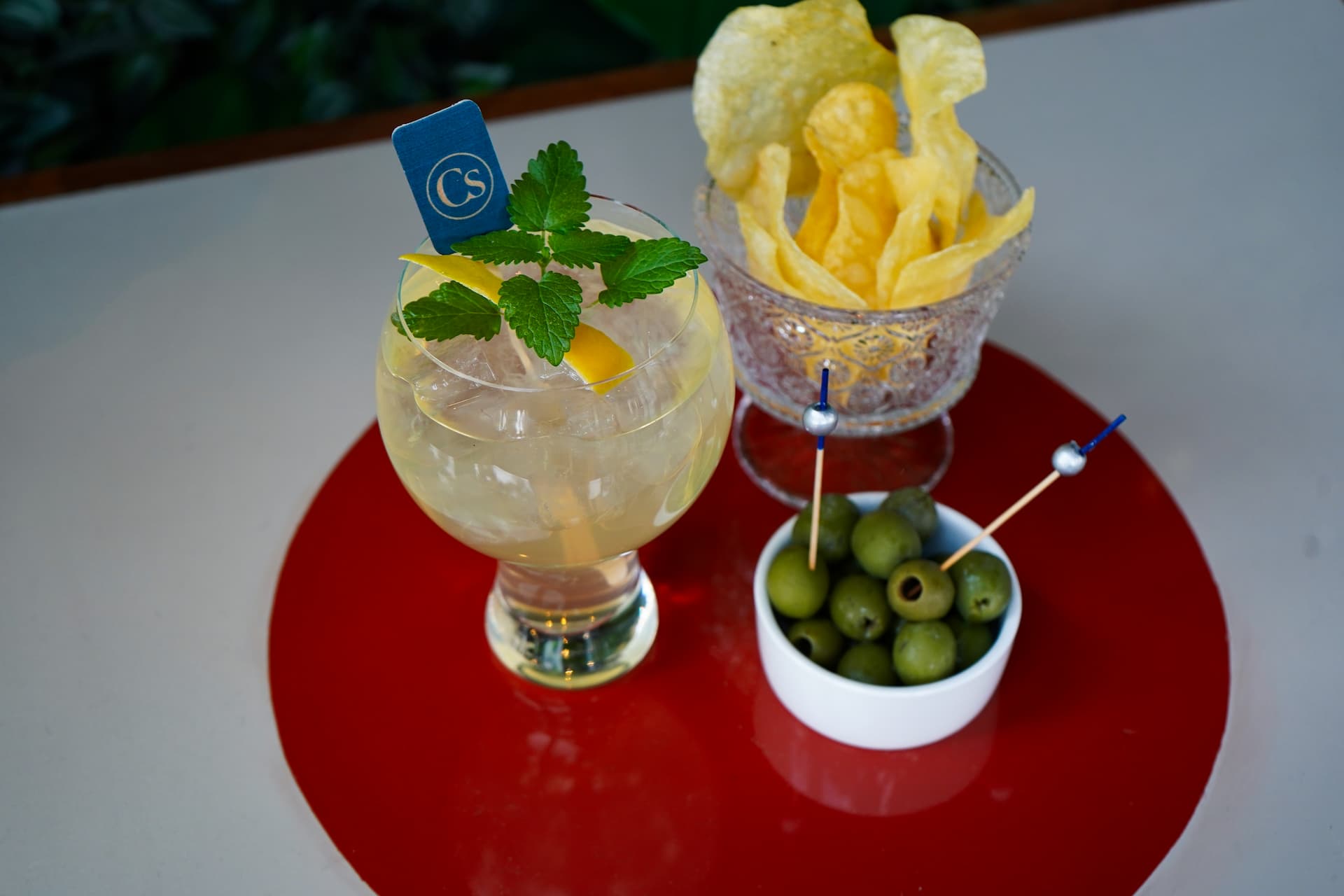 Light-colored cocktail garnished with fresh mint and lemon peel, served with green olives and potato chips on a red tabletop.