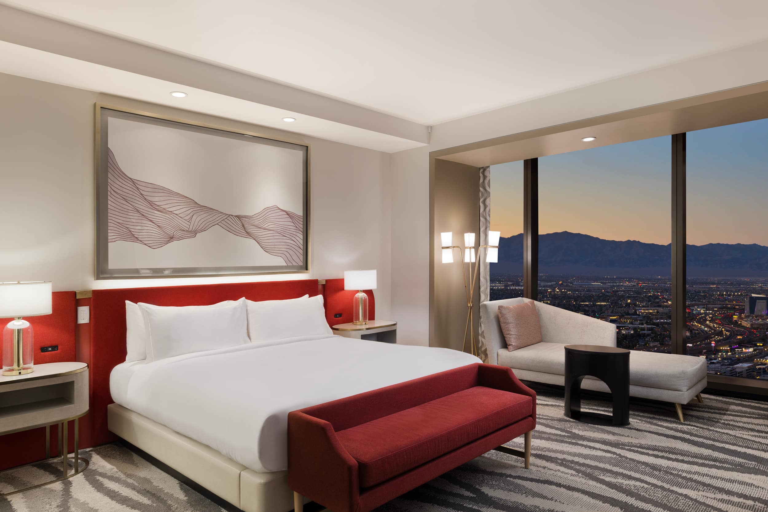 A luxury hotel bedroom with a white king bed, vibrant red headboard and matching bench, a large framed wave-line art piece, and a chaise lounge sitting in front of a floor-to-ceiling window overlooking a city and mountain sunset.