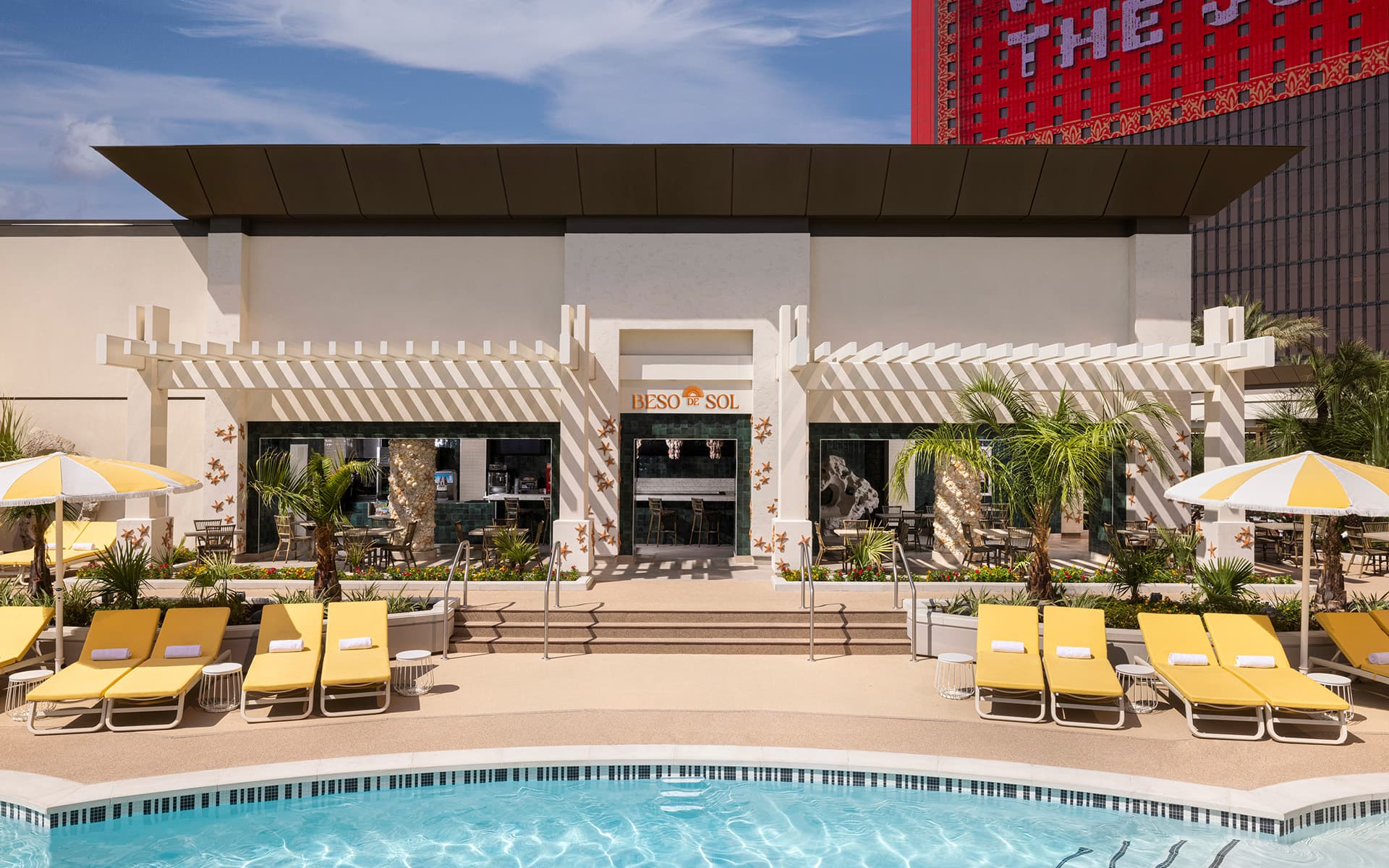 Exterior view of Beso del Sol poolside restaurant at Resorts World Las Vegas, featuring yellow lounge chairs, striped umbrellas, palm trees, outdoor seating, and the resort pool in the foreground.