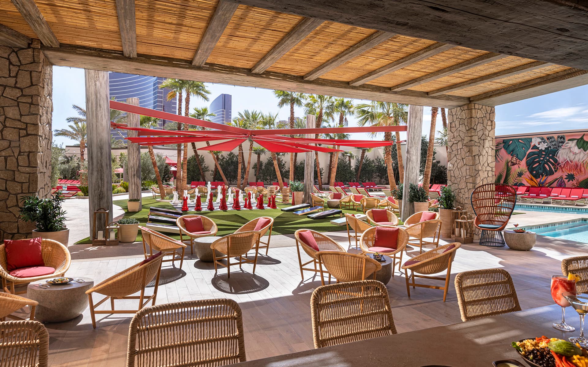 Open-air view of the Bimini Pool Bar at Resorts World Las Vegas featuring wicker lounge seating, shaded dining areas, oversized outdoor games, palm trees, and a resort pool with red lounge chairs in the background.