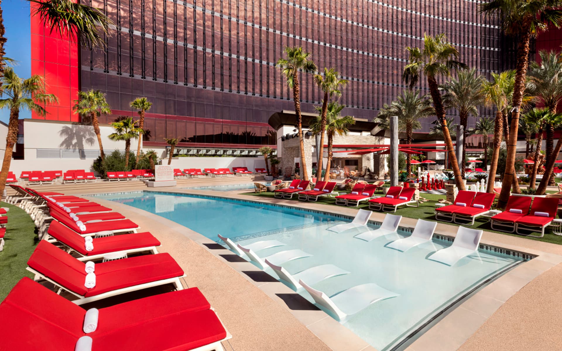 Wide daytime view of the Bimini Pool featuring crystal-clear water, in-water lounge chairs, rows of red cushioned loungers, palm trees, and the Resorts World Las Vegas hotel tower in the background.