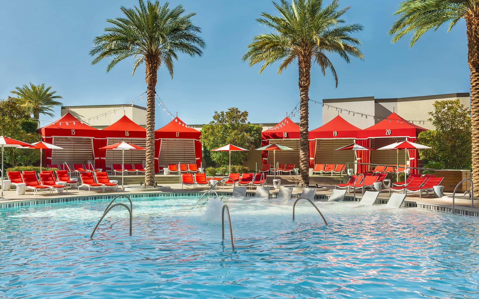 View of the Crockfords pool at Resorts World Las Vegas featuring turquoise water, in-water loungers, red cabanas, palm trees, and poolside seating under a clear blue sky.