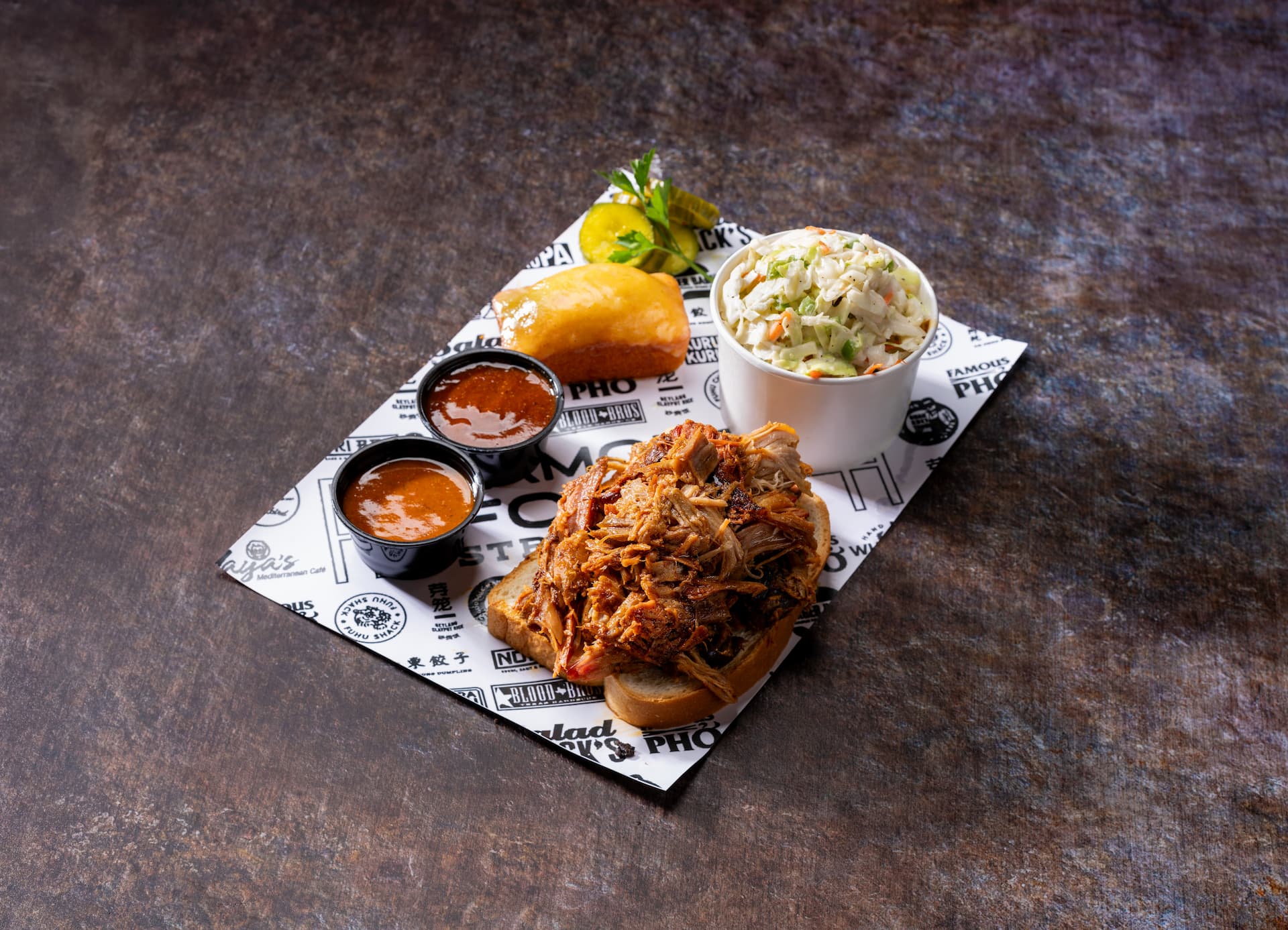 Pulled pork served on toasted bread with barbecue sauce, coleslaw, pickles, and a dinner roll on a Famous Foods serving tray.