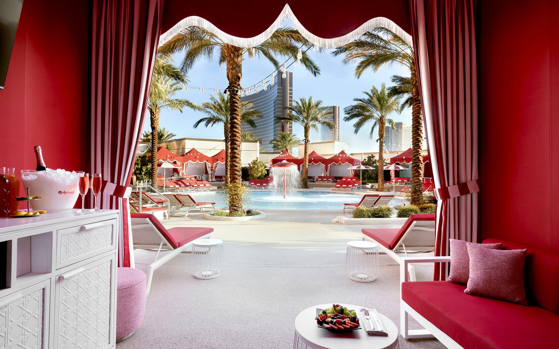 Luxury poolside cabana with red seating and curtains overlooking a resort pool, palm trees, private cabanas, and the Las Vegas skyline on a sunny day.