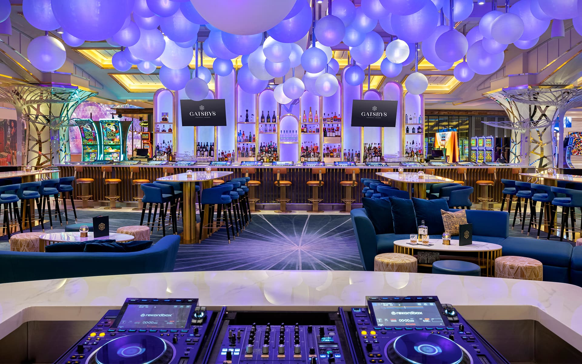 Interior of Gatsby’s Cocktail Lounge at Resorts World Las Vegas featuring a central bar, blue lounge seating, illuminated globe fixtures, and a DJ booth.
